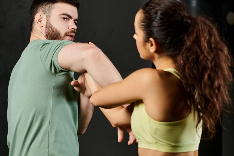 A male trainer teaches self-defense techniques to a woman in a gym setting.